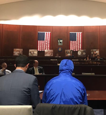 a man in a blue hooded jacked next to a man in a suit, both photographed from behind in a committee room