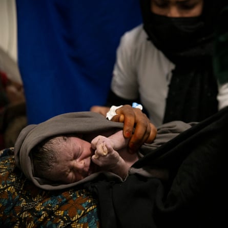 A midwife wearing a hijab holds a newborn baby wrapped in a blanket