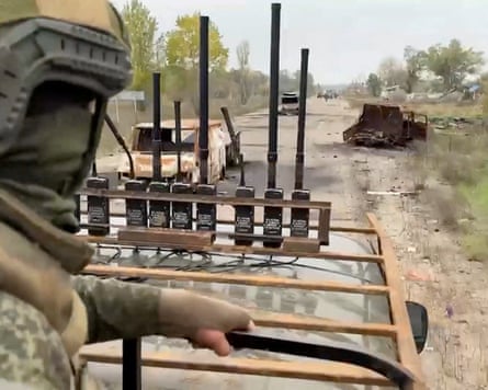 A in a military vehicle, a Russian soldier drives past burnt-out, rusted vehicles abandoned at the side of a road
