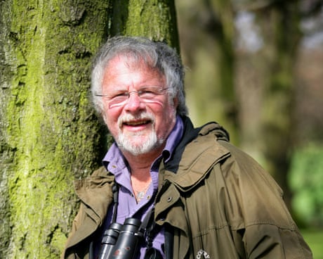 He smiles and leans against a tree, wearing binoculars