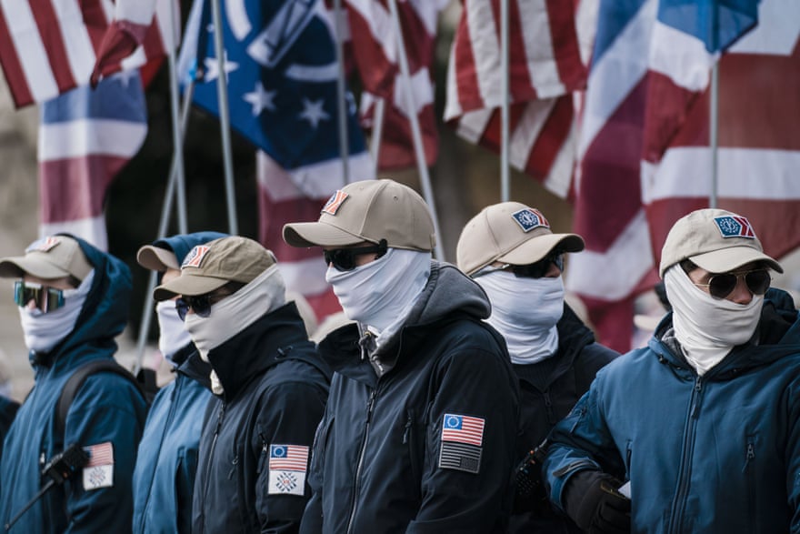 A group of men wearing balaclavas covering their faces and khaki colored caps stand in front of a row of American flags