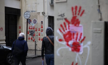 Graffiti depicting red handprints are seen painted on walls in the street; a cluster of three are seen close-up on one side of the road and then more on a larger space by a doorway on the other, among stickers and other images. Two people are seen from behind crossing the road; the street is wet and it looks as if it is raining.