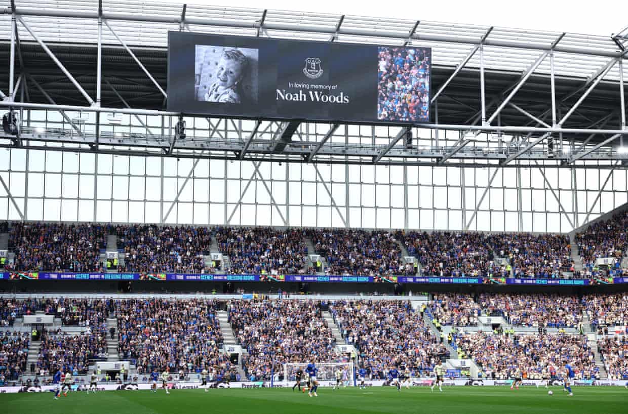 The interior of a football stadium with a big screen, stand and players on the pitch all visible.