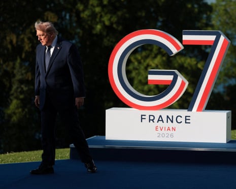 Donald Trump stands by a G7 sign in the red, white and blue of the French flag. He is facing to one side and his hair is blowing in the breeze.