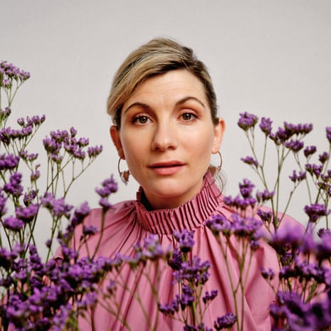 A portrait of Jodie Whittaker wearing a pink dress with a ruffled collar, behind some purple flowers