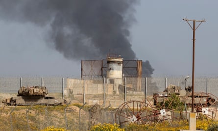 Black smoke rises behind the border fence; armoured vehicles and what may be a watchtower and agricultural equipment are in the foreground
