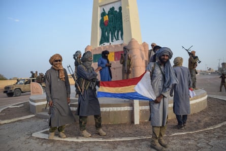 FLA fighters stand around a monument on a roundabout holding a red, gold, blue and white flag. They wear loose tunics over their khaki and camouflage trousers and have scarves wrapped around their heads. They carry guns over their shoulders.