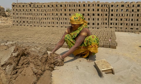 An Indian woman squats down beside a big pile of clay and a mould, with stacks of finished bricks behind her