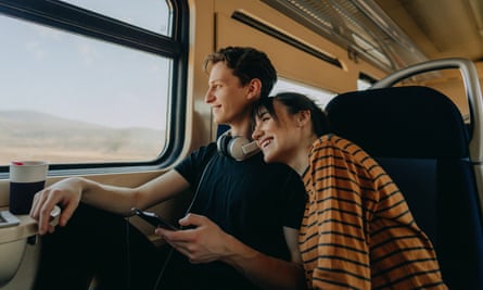 a smiling young couple travelling together by train in Serbia – they are sitting by the window, he has a dark T-shirt and is wearing headphones around his neck, while she has an orange T-shirt with black stripes; she is resting her head on his shoulder