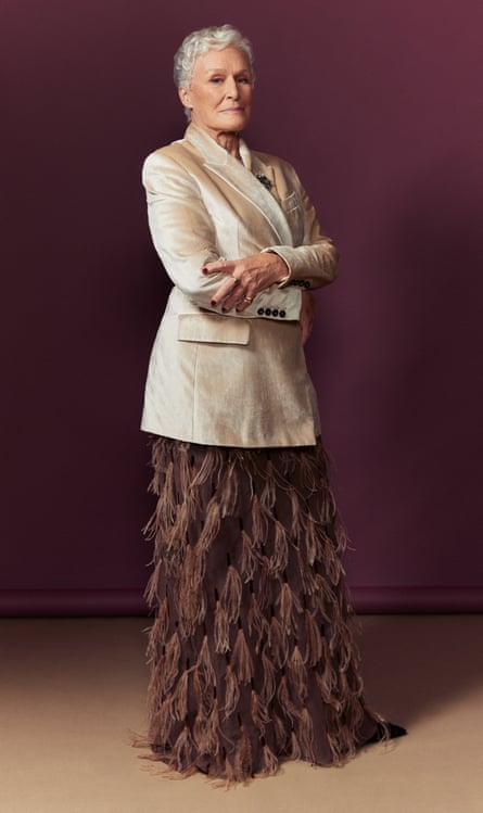 Glenn Close standing at an angle to the camera with her arms folded, wearing a smart cream blazer and full-length brown feathered skirt