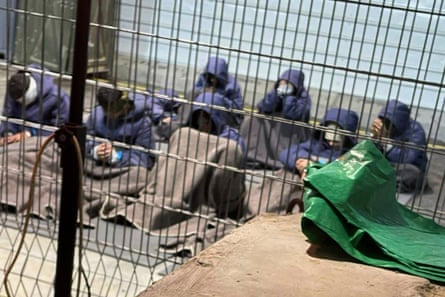 Prisoners with their hands and legs restrained sit on the floor of a cell, wearing blue uniforms with hoods up