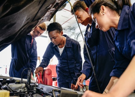 A female apprentice attaches electrical jump leads to an engine with a car bonnet raised, with her fellow students watching. They are all wearing dark blue overalls.