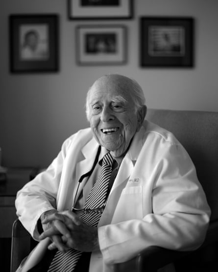 A black and white photograph of an elderly man seated in a chair. He is wearing a doctor’s lab coat and is smiling widely. His hands are clasped in front of him.