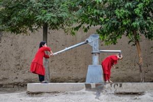 Fayzabad, Afghanistan. A girl in red has her mouth next to the water outlet of a metal pump as another girl, also dressed in red, works the force rod