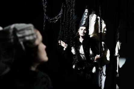 Abby McCann in a black cone hat labeled “Rumour” stands amid hanging chains, facing Sophie Kean