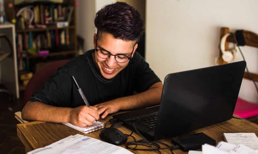 Young man at home working on his personal finances. He is sitting at a wooden table with a black laptop and bills, doing some paper work. He uses glasses and a black t-shirt.