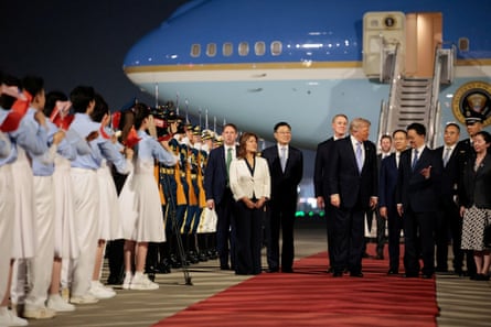 Donald Trump stands on a red carpet with Han Zheng in front of the US Air Force One plane. A line of young people in blue and white costumes wave red flags.