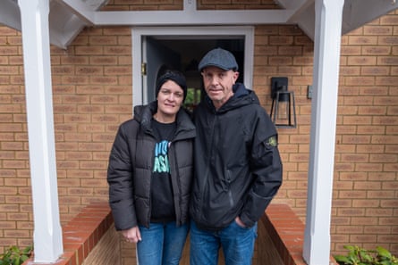 James and Faye Wade at the front of their modern brick house; they are standing in a porch with white pillars. Both wear blue jeans and black anoraks.