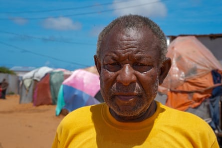 An older Somali man stands in front of his small crude shelters of sticks, cloth and plastic
