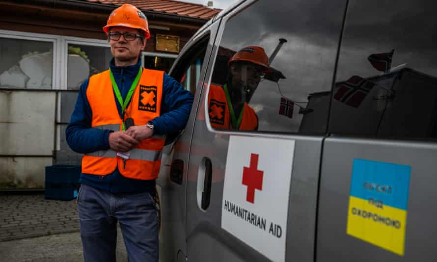Co-founder of Paracrew Kristoffer Rolf Deinoff wears an orange hi-vis tabard and is leaning against a van with a Ukrainian flag sticker on the door and a sticker with a red cross and the words ‘humanitarian aid’.