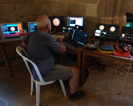 A man sits at a workstation in a stone-walled room, surrounded by multiple computer monitors displaying images of the Sun and astronomical data.
