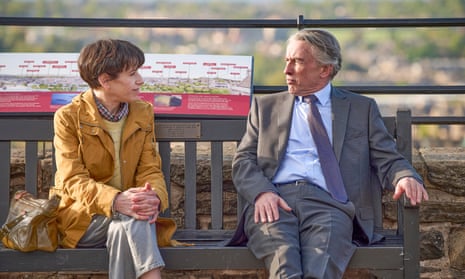 Sally Hawkins as Philippa Langley with Steve Coogan as John Langley in The Lost King: they are sitting on a wooden bench in front of a historical site. Hawkins wears a mustard-yellow anorak and Coogan a grey suit and tie, dressed as their characters.