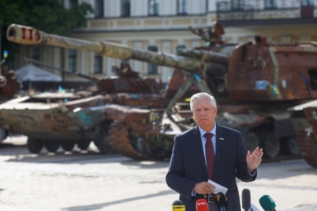 a man speaks into microphones in front of a tank