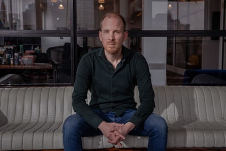 he sits on a pale, low sofa leaning forward with his forearms across his legs and knees apart. He is tall and thin with short, reddish-blond hair and beard, and wears a dark green shirt and blue jeans.