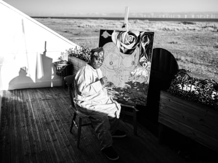 O Yemi Tubi sits outside wearing a hat and robe with one of his paintings