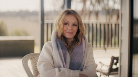 Kylie Minogue sitting on a porch smiling and wearing knitted clothes