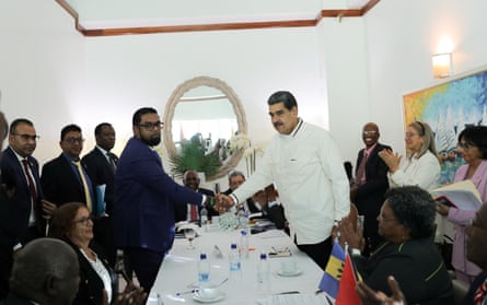 Two men shake hands across a table as their entourages applaud
