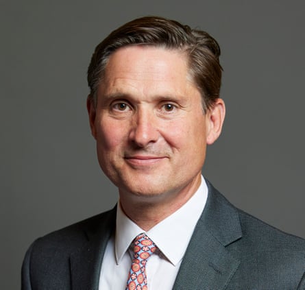 Charlie Maynard smiles slightly in a formal portrait, wearing a grey suit