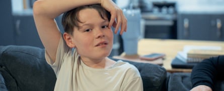 Mason sits with one hand over his head. He has light brown hair with a side-parted fringe over his forehead and wears a white T-shirt.