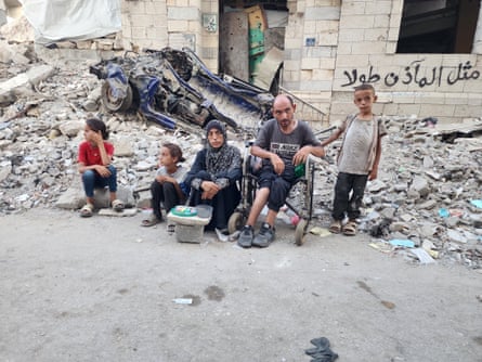 The Khalidi family sit in front of the rubble of a bombed mosque while in front of them is a table where children sell bracelets made of beads.