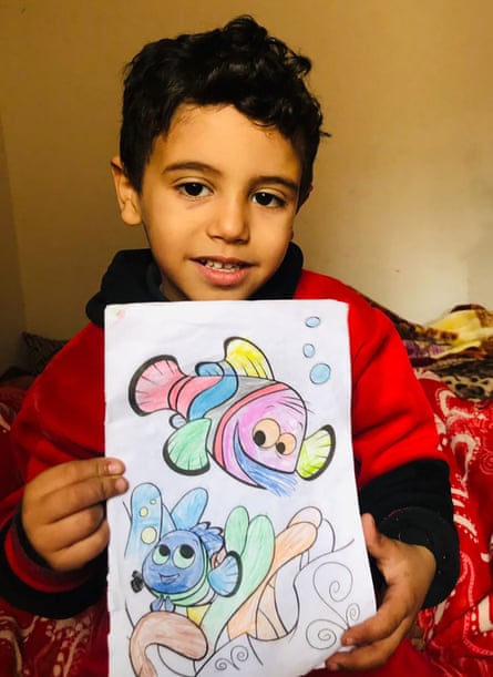 A young boy in a red jumper looks at the viewer, holding up a picture of two cartoon fish that have been coloured in with crayons.