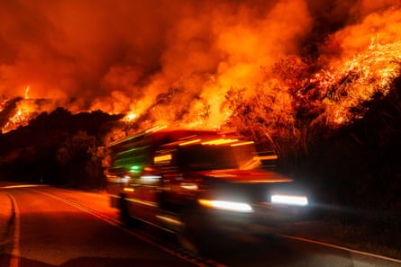 a fire truck drives along a road as a fire rages