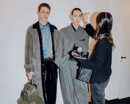 Two models wearing grey Armani clothes have makeup applied backstage.