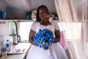 Memory poses inside a caravan wearing a wedding dress with lace bodice and holding a blue bouquet of flowers