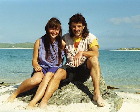 Steel and Dines sit together on a sandy beach with turquoise water and islands behind them