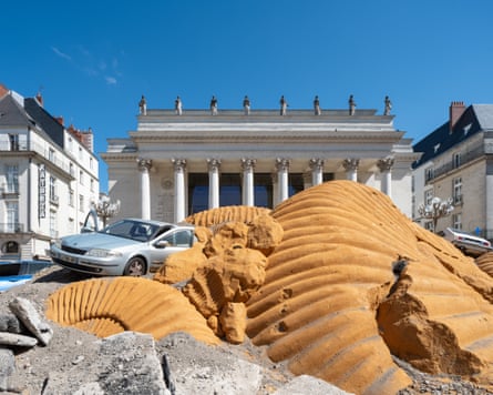 A mound of large ammonite sculptures, rubble and an abandoned car in front of a neoclassical theatre