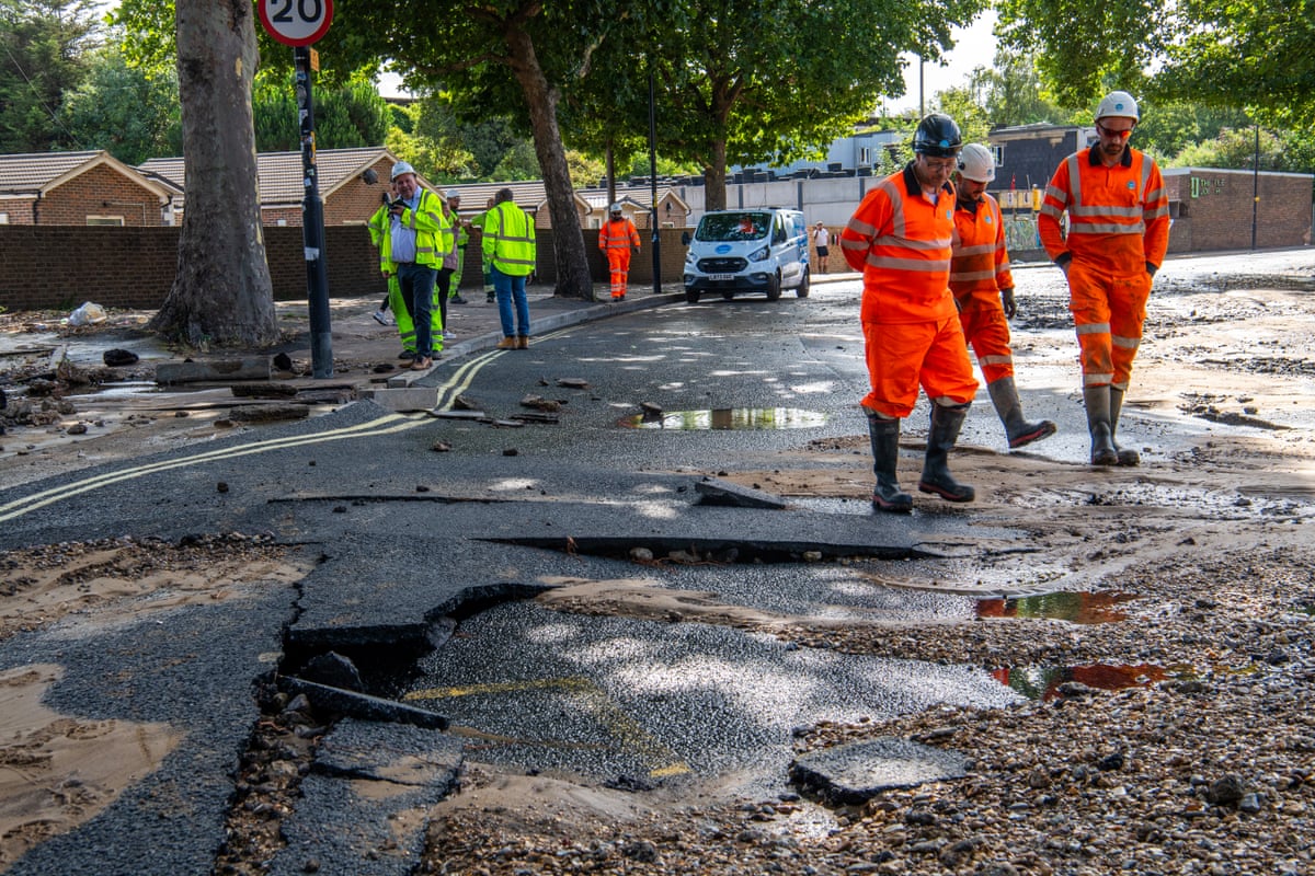 Men in orange hi-vis overalls and wellies stand by puddles in a road where the surface is cracked and potholed. Others in yellow vests stand in the background.