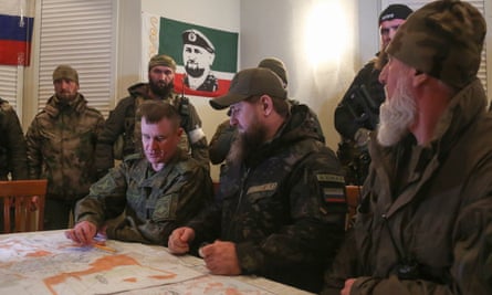 Ramzan Kadyrov sitting at a table with a map on it alongside a Russian officer while surrounded by armed soldiers