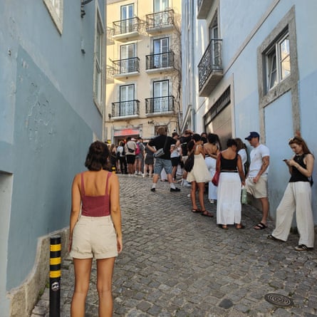 A woman in shorts stands with her back to the camera in a cobbled street looking at a queue of tourists