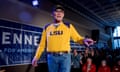 An older white man wearing a ball cap, glasses and bright yellow and purple LSU shirt and jeans, speaks and points on a small stage.