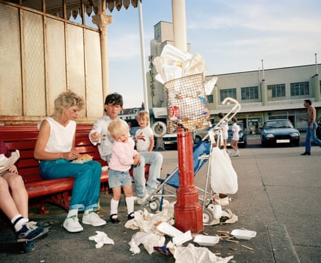 A family sitting on a bench in a British seaside resort in the early 1980s, an overflowing rubbish bin in front of them