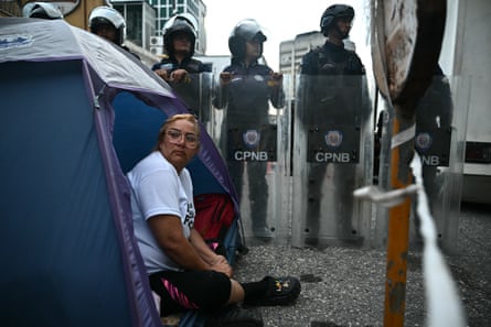 a woman sits in a tent next to a line of riot police