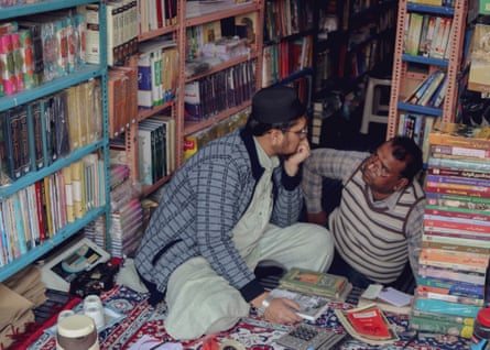 A man seated amid stacks and shelves of books leans his face on his hand and talks to an older man.