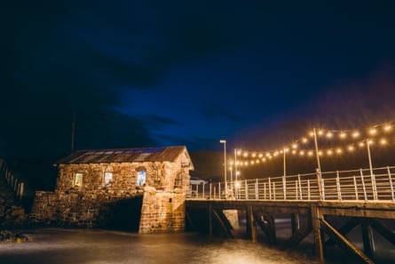 A stone building with a corrugated roof by the sea in north-west Scotland at night, with lights on within.