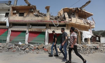Three young men walk along a rubble-strewn street past a low two-storey building, the upper floor of which has been completely destroyed
