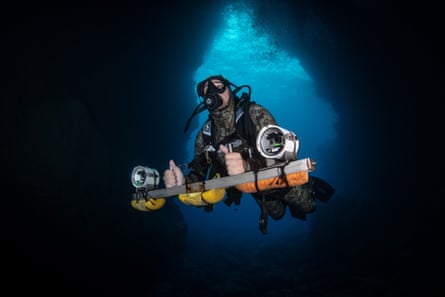 A man in a camouflage wetsuit, with a scuba tank and two large underwater cameras mounted on a metal bar in his hands, swims into a cave